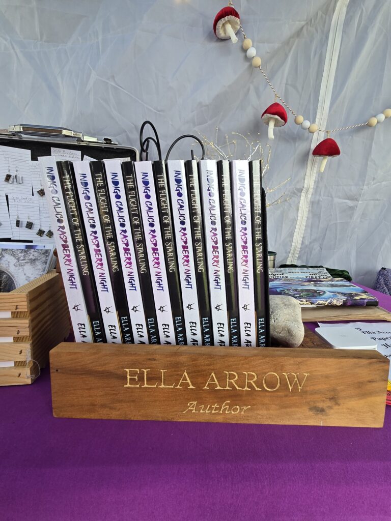 Books by Ella Arrow behind a wooden sign, "Ella Arrow, Author"