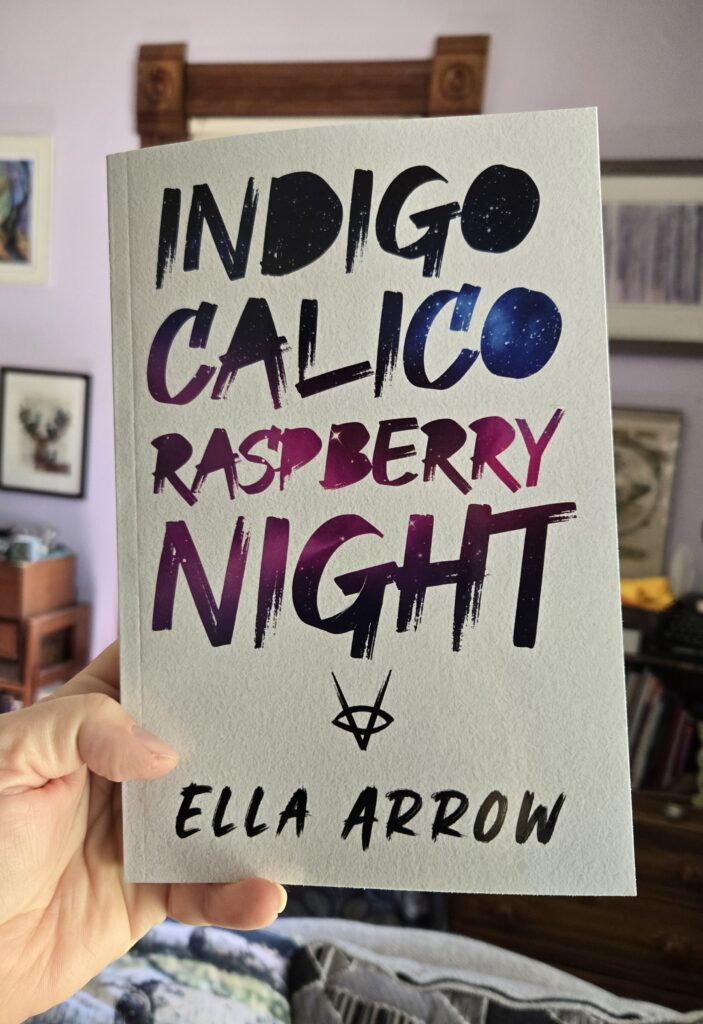 Hand holding up paperback book, Indigo Calico Raspberry Night by Ella Arrow