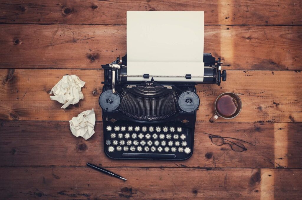 Typewriter on a wooden table with coffee and crumpled pages. New YA fantasy book by Ella Arrow was not written on a this typewriter.